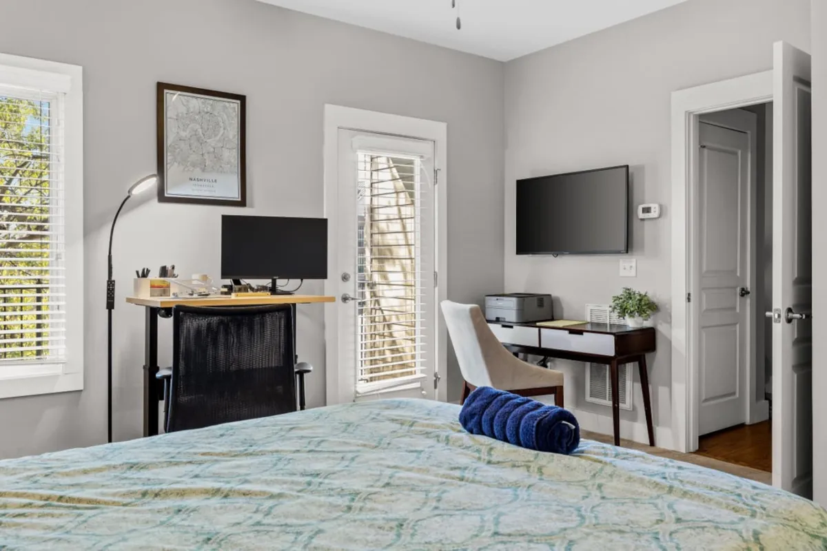 A comfortable bedroom setup with a cozy bed adorned with a decorative blanket, a desk set up for work or study, and a large window letting in natural light. The space feels both relaxing and functional, catering to various guest needs.
