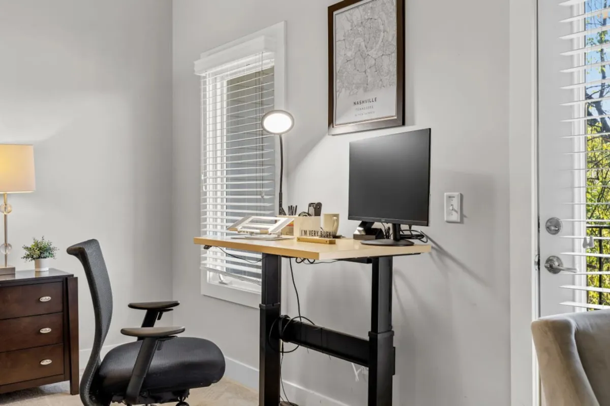 A well-lit workspace featuring a standing desk equipped with a computer monitor and desktop lamp. The office area includes a framed map on the wall and a nightstand with a lamp, evoking a productive yet comfortable atmosphere ideal for work or study. The decor is clean and professional, with a focus on functionality and comfort.