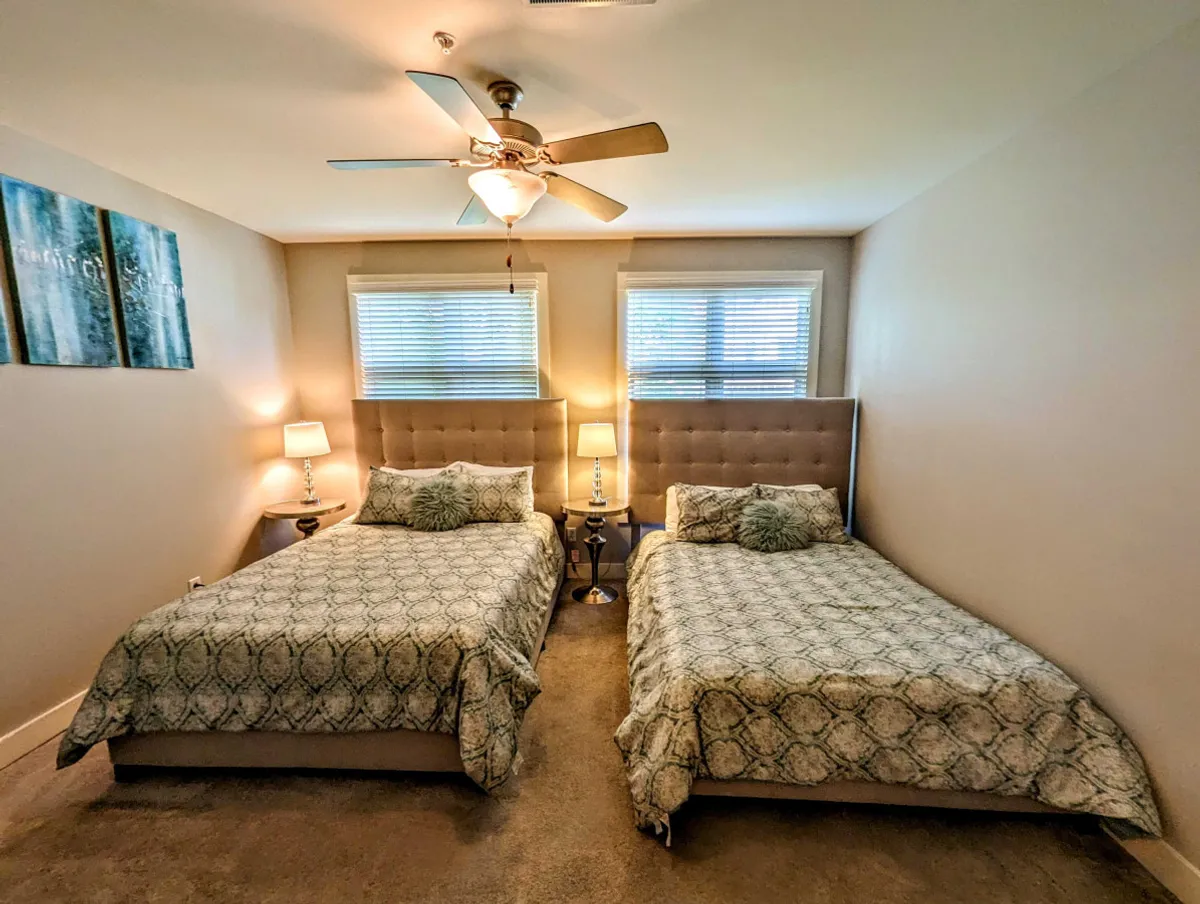 This image showcases a serene bedroom with two beds featuring patterned bedding, elegant nightstands with matching lamps, and stylish decorative wall art. The beds are arranged to maximize space and comfort, while sunlight softly brightens the room, creating an inviting atmosphere.