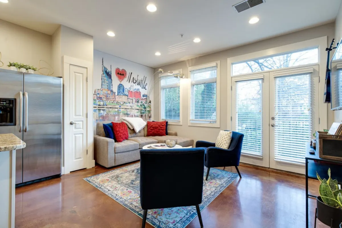 A cozy living room featuring a plush sofa, artfully arranged wall decor, a modern coffee table, and large windows allowing abundant natural light. The layout suggests an inviting space for relaxation and social gatherings.