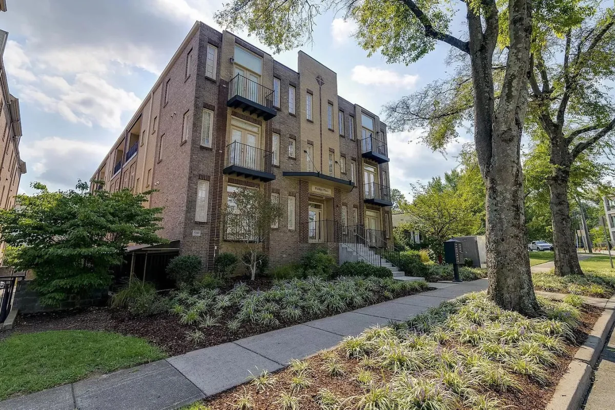 This image features the brick exterior of the property with welcoming balconies, a landscaped front yard, and a well-maintained sidewalk. The setting feels inviting, suggesting a walkable, vibrant neighborhood just outside the front door.