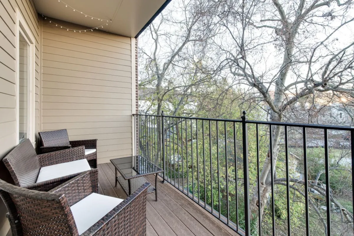 An outdoor balcony furnished with wicker chairs and a glass table overlooks a view of leafy trees. The space exudes a calm and inviting atmosphere, ideal for relaxing after exploring the city.