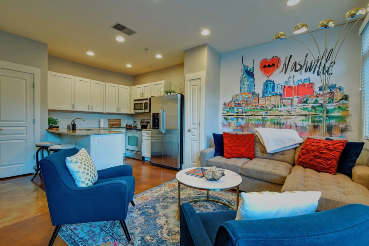 The image showcases a stylish, open-concept living area featuring a modern kitchen, cozy sofa, vibrant local art on the walls, and a welcoming coffee table—perfect for group gatherings. The decor subtly nods to Nashville's music scene, and the setup invites both relaxation and socializing.